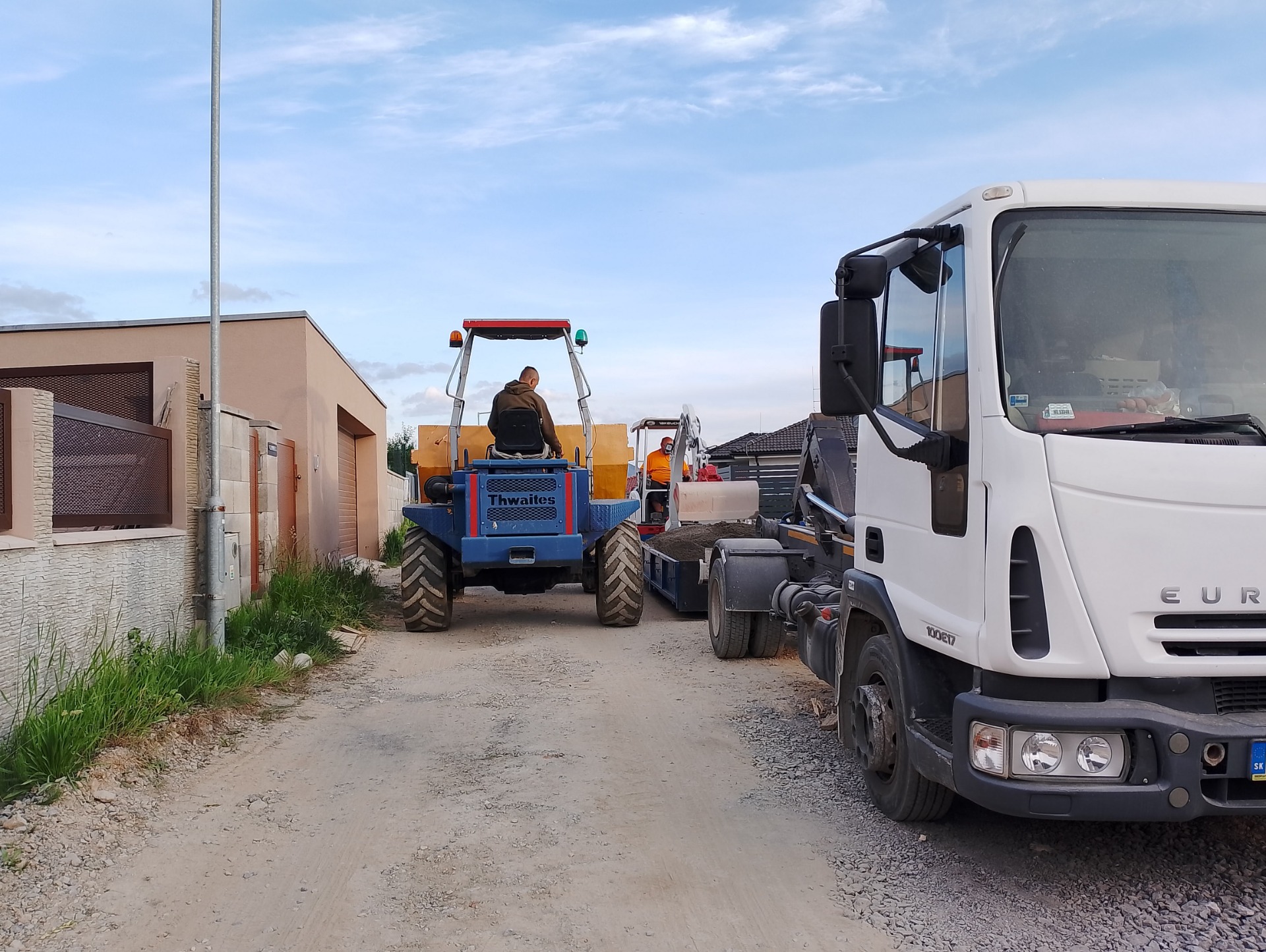 Construction worker drives a blue telehandler in a yard while a white truck waits nearby.