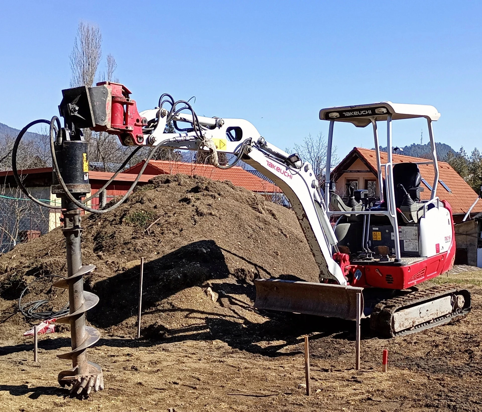 Red and white excavator drilling into soil, with hydraulic auger and houses in background under blue sky.