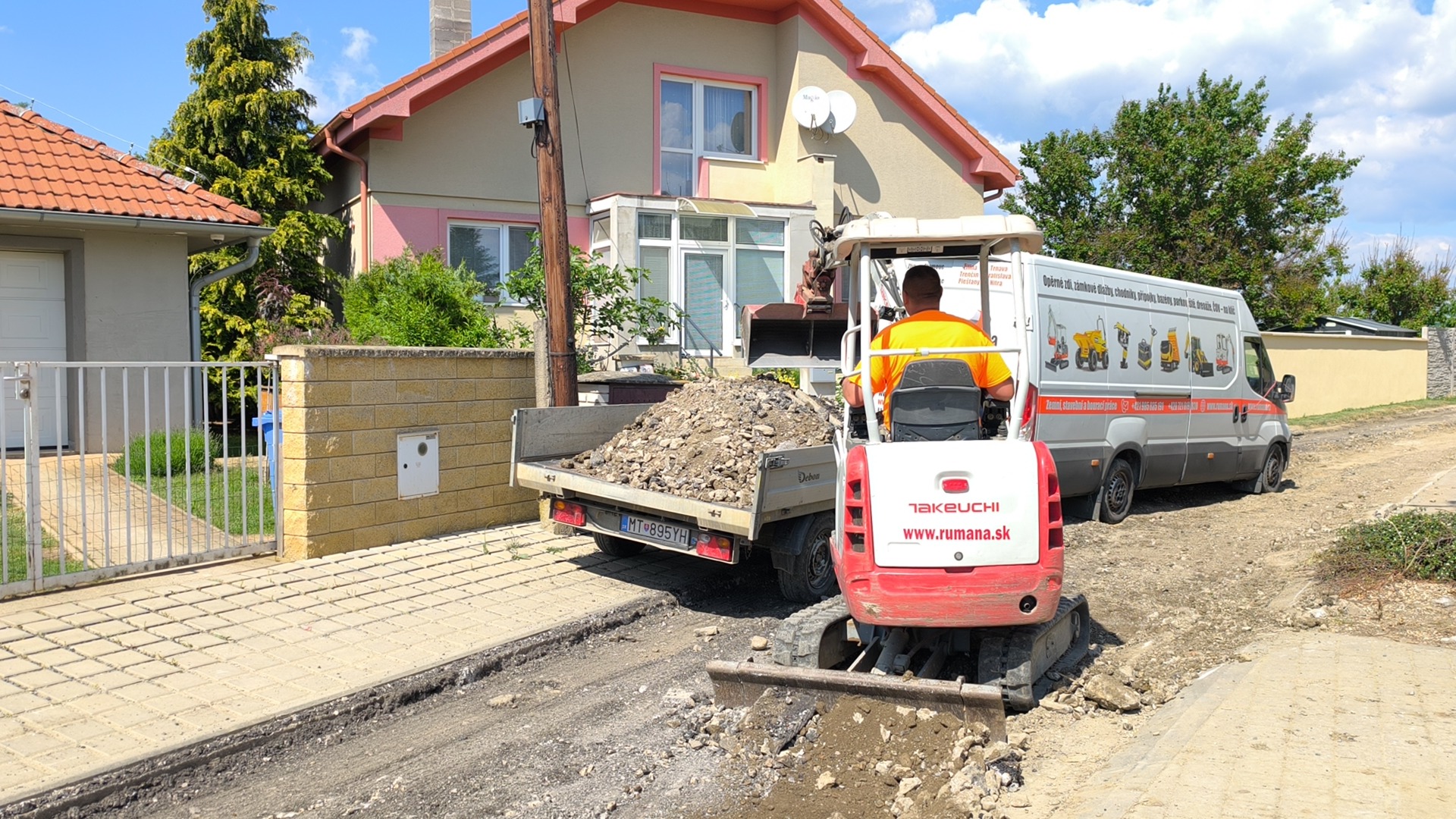 Červený bagr na přívěsu nakládá suti u rodinného domu, vedle stojí pracovník v oranžové vestě.