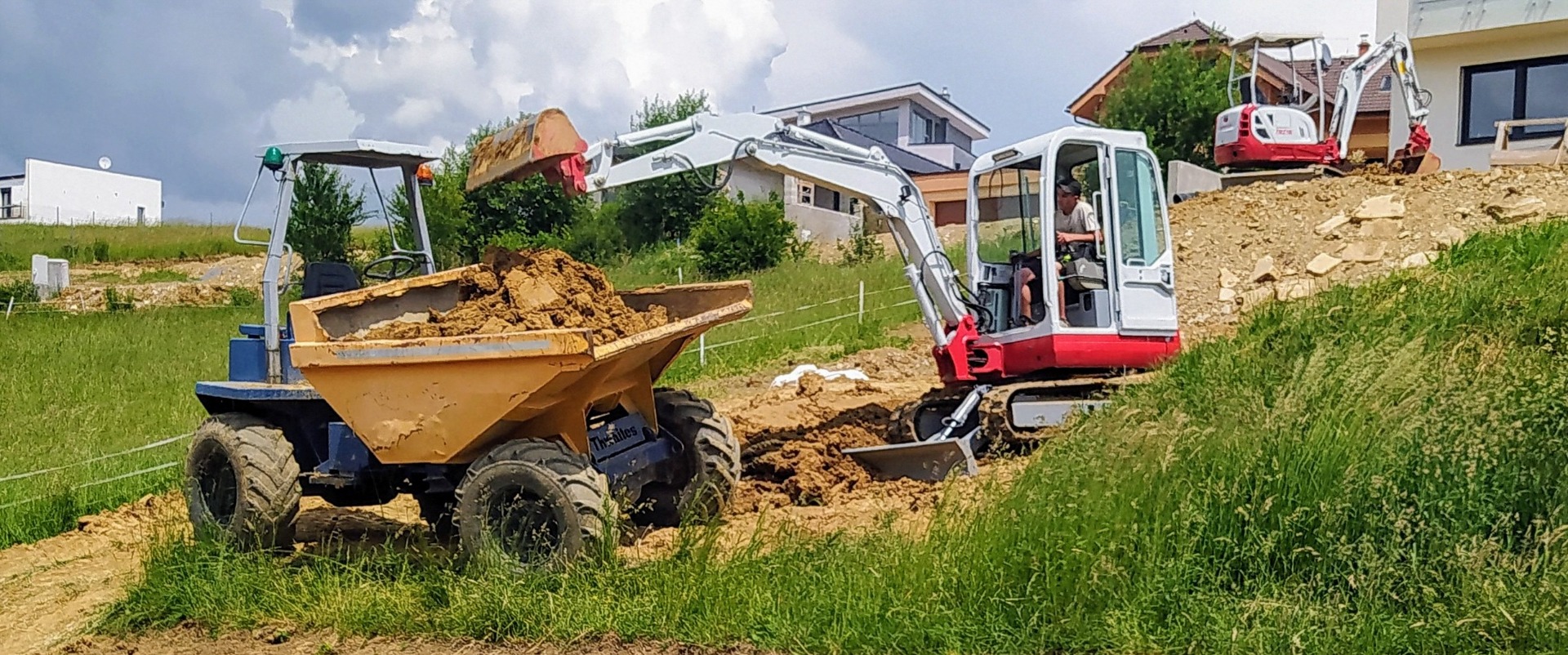 Construction excavator and dump truck moving soil on a grassy hillside near houses under a cloudy sky.