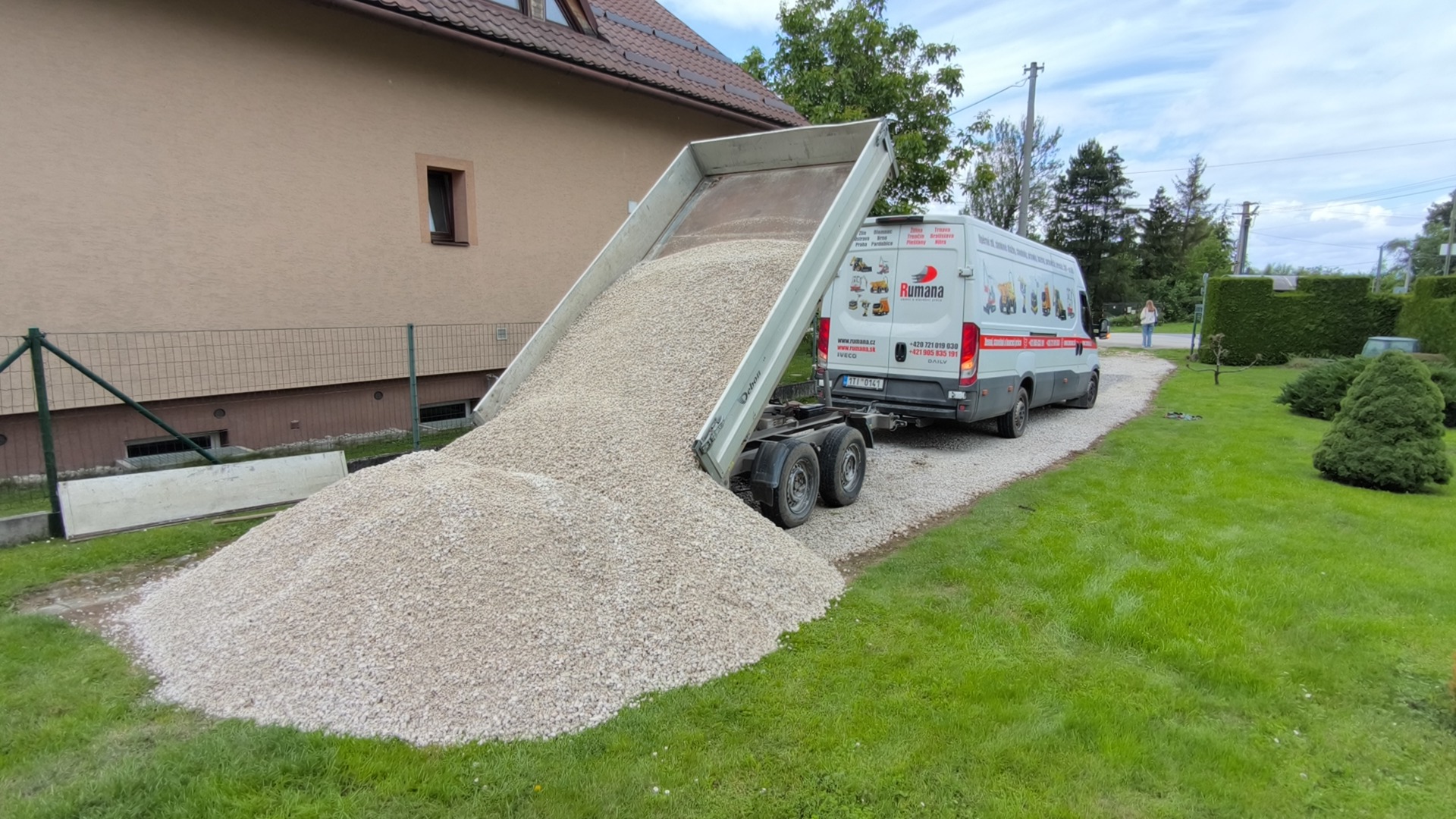 Nákladní vůz s otevřenou sklápěčkou vysypává štěrk na štěrkovou plochu u rodinného domu.