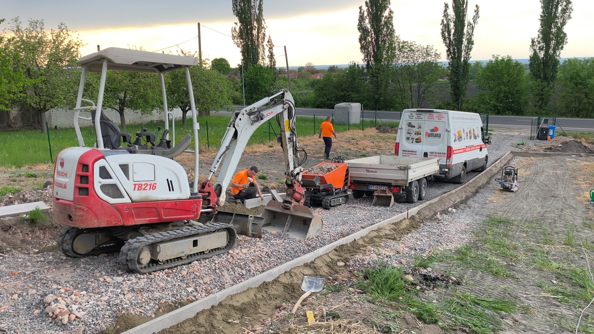 Stavební bagr s ramenem nakládá štěrk, pracovníci pracují u nákladního auta na stavbě.