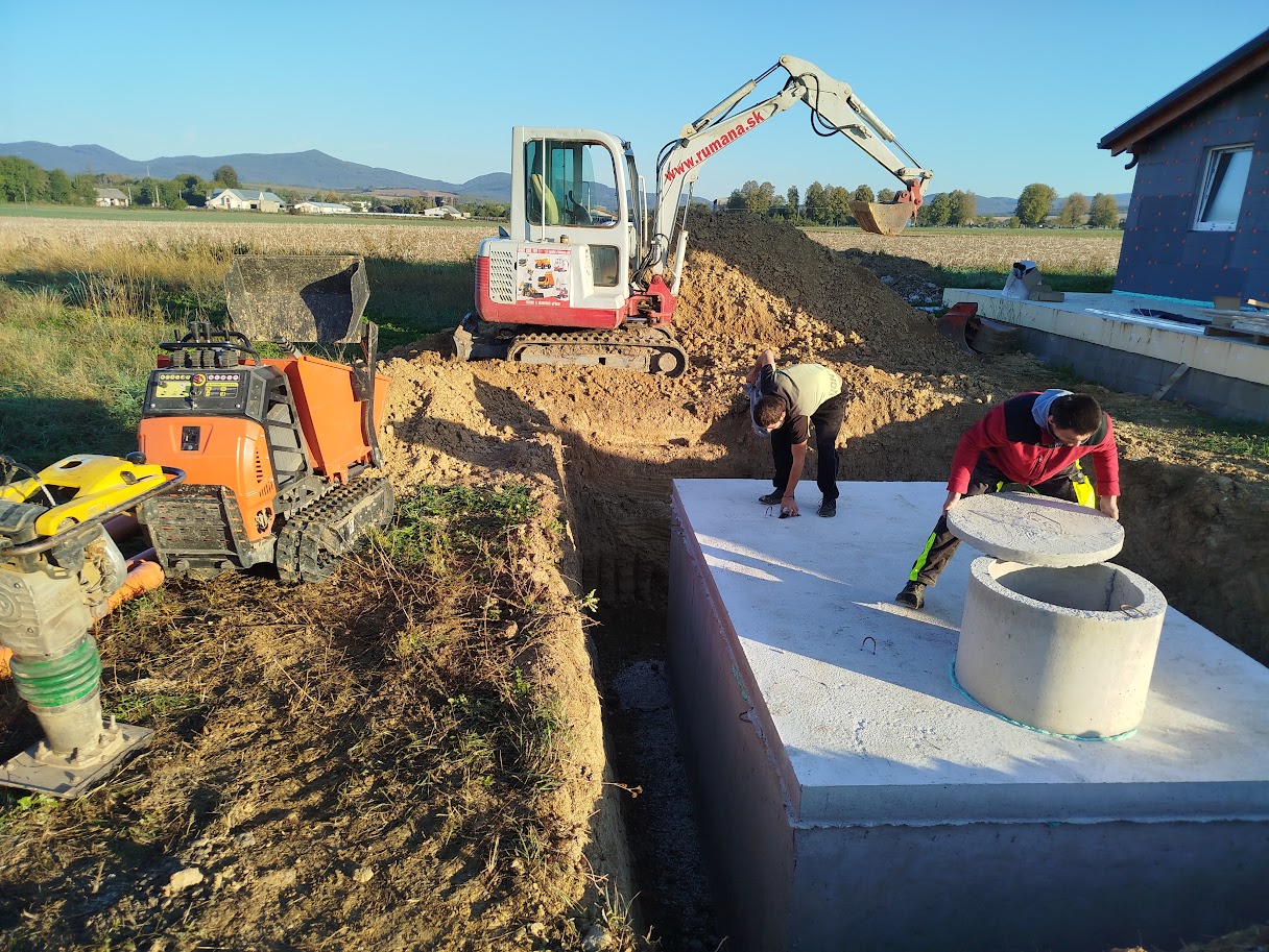 Bagrovací stroj a pracovníci při výkopu a pokládce betonové desky na venkovském staveništi.